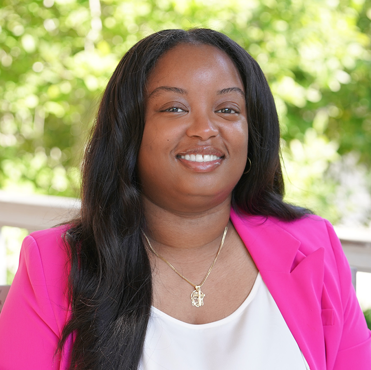 Smiling woman in a vibrant pink blazer, outdoors with green foliage.