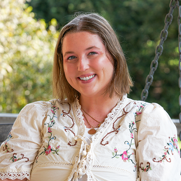 Young woman with light dark blonde hair smiles, wearing an embroidered cream top.